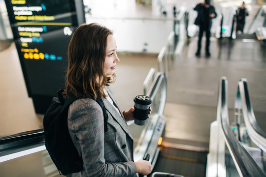 Young pretty girl with a backpack going down the escalator at the airpot, holding black coffee cup.