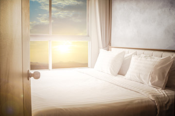 Open Door to bed in hotel room bedroom white bed sheets lit by natural warm nature sunlight from window at morning time
