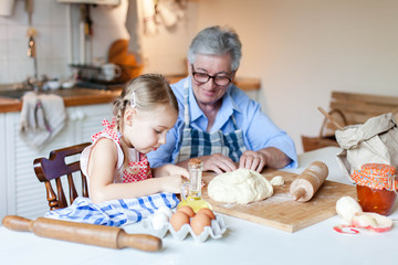 Grandmother is teaching child to cook pastries and apple pie in cozy home kitchen. Senior woman and little girl are happy together. Cute kid is helping to prepare dough. Lifestyle authentic moments.