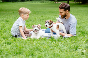 Obraz premium Beard man cynologist with little boy son lying playing relaxing on a green grass with two dogs at green park summer time. Family, pet, team, friendship domestic animal and people concept