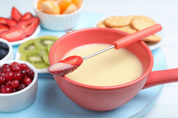 Fork with strawberry dipped into white chocolate fondue and ceramic pot on table