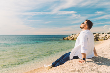 Naklejka na ściany i meble young stylish man sitting at sea beach enjoying the view