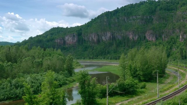 Aerial approach shot.Crossing the railroad,electic power lines and follow the river towards rocky mountains.The camera flies close on top of the tree tips.