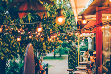 Friendly youth atmosphere in a cozy outdoor summer terrace in a coffee shop. Street garland decorates the space. Defocused image to convey the mood, blurred background.