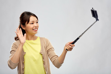 technology and people concept - happy asian woman taking picture by smartphone on selfie stick and waving hand over grey background