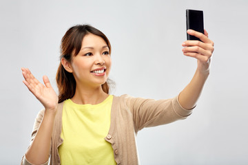 technology and people concept - happy asian woman taking selfie by smartphone and waving hand over grey background