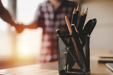 pencil and tools in pen holder stand on desk with construction worker team hands shaking greeting...