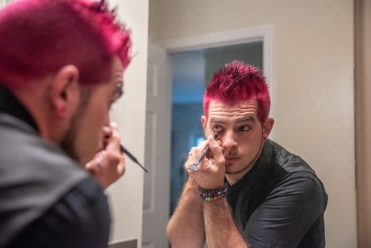 Diverse caucasian man with spiked pink hair applying eyeliner in the mirror