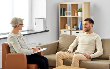 psychology, mental therapy and people concept - smiling senior woman psychologist with clipboard talking to young man patient at psychotherapy session - obrazy, fototapety, plakaty