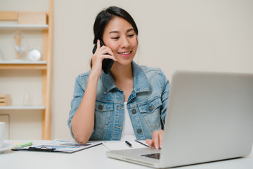 Beautiful smart business Asian woman in smart casual wear working on laptop and talking on phone while sitting on table in creative office. Lifestyle women working at home concept.