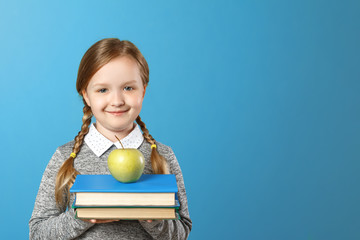 Portrait of a cheerful little girl on a blue background. Schoolgirl is holding a stack of books and an apple. Back to school. The concept of education.