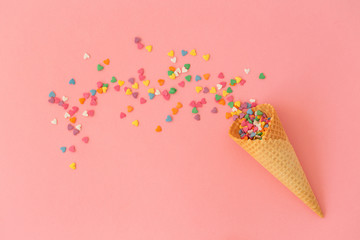 Empty ice-cream waffle cones and colorful sprinkles on pink background, top view. Scattering of multicolored sweets and confectionery topping heart shaped. 