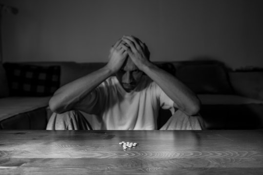 Depressed man suffering from suicidal depression want to commit suicide by overdose strong medicament drugs and pills pain killers while he sitting in dark room selective focus moody dramatic look