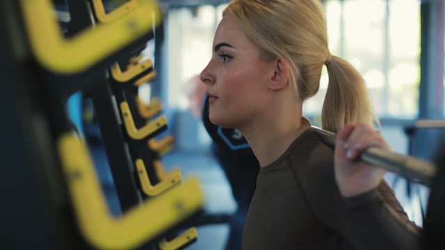 beautiful lady with fair hair squats with silver color barbell against man silhouette in gym closeup slow motion