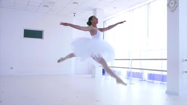 Ballet dancer is jumping up making grand temps leve passe. Classical dance in white tutu on pointe. She is performancing splits in the air. Young ballerina in light class with big windows.