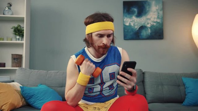 Portrait of concentrated retro fun weak man training with light dumbbells at home. Funny vlogger in colorful sports outfit doing exercises on smartphone camera.