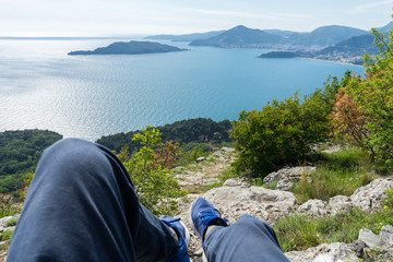 First person perspective sightseeing of adriatic coast in Budva, Montenegro. Legs with blue sneakers of a traveler in a cliff with green mountain and blue sea and islands trail