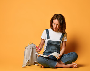 girl in a white T-shirt and denim overalls, sitting on the floor with a red notebook and a pencil on a yellow background