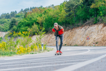 Obraz premium Tall fit male longboarder riding his longboard downhill on a sunny day high in nature while wearing a red shirt, green hat, and grey jeans.