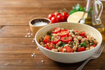 White bean salad with tomatoes, capers, garlic and parsley in bowl on wooden table. Selective focus.