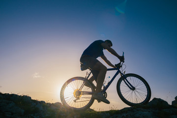 Naklejka na ściany i meble Silhouette of a fit male mountain biker riding his bike uphill on rocky harsh terrain on a sunset.