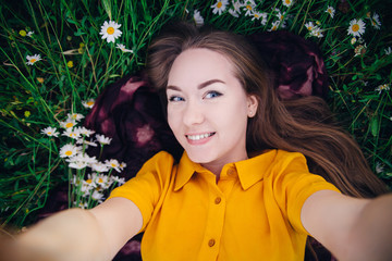 Cute girl in a bright yellow dress lies among a flower field and makes a selfie