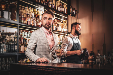 Handsome modern man in checkered suit is consuming alcohol at trendy bar.