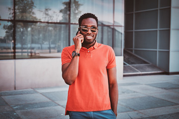 Portrait of cheerful african student with sunglasses and mobile phone near glass building.