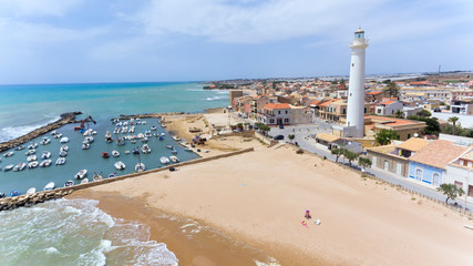 Golden sandy beach with shallow waters, small port with boats, white tall lighthouse in Sicilian fishing village of Punta Secca, set of TV series Inspector Montalbano, aerial view . - 275171695