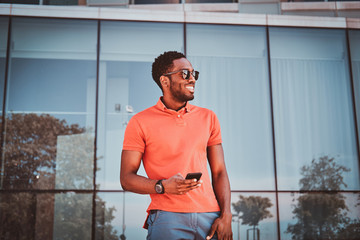 Portrait of cheerful african student with sunglasses and mobile phone near glass building.