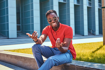 Obraz premium Young happy afro american man in sunglasses is sitting on the grass near skyscraper.