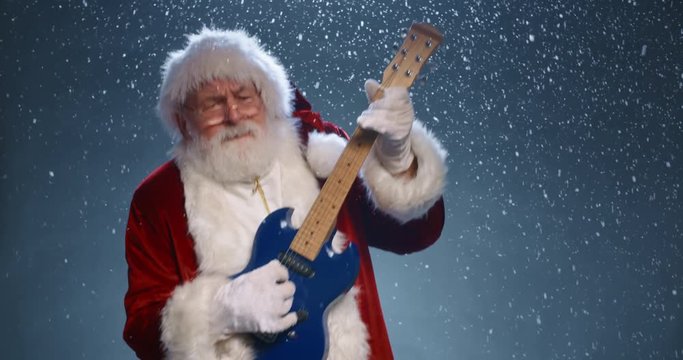 Funny santa clause is playing electric guitar and dancing while looking at camera, isolated over snowy blue background - christmas party, christmas spirit concept close up 4k footage