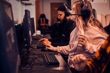 Focused girl get exited about videogame. She is wearing head set and glasses. another gamers at background.