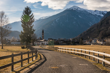 Road fenced with a wooden fence leading to alpine village. Cloudy sky and snow-capped mountains Italian Alps at the background. Trento, Italy.