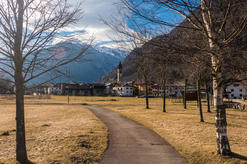 Pathway between trees leading to alpine village. Cloudy sky and snow-capped mountains Italian Alps at the background. Pellizzano, Trento, Italy.