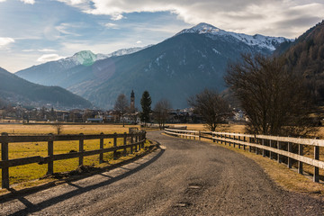 Road fenced with a wooden fence leading to alpine village. Cloudy sky and snow-capped mountains Italian Alps at the background. Trento, Italy.