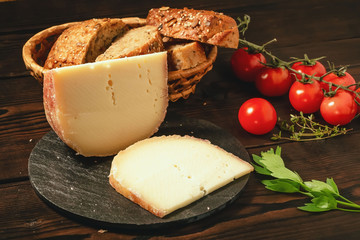 Pieces of hard cheese on slate dish, parsley, thyme, grain bread and cherry tomatoes on a wooden table