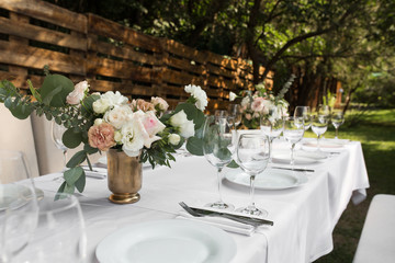 Wedding table setting decorated with fresh flowers in a brass vase. Wedding floristry. Banquet table for guests outdoors with a view of green nature. Bouquet with roses, eustoma and eucalyptus leaves