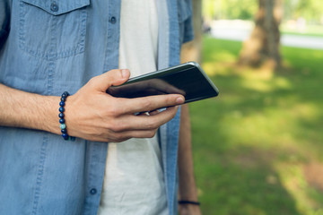 Close up on midsection of young man holding mobile smart phone in his hand in park in summer day