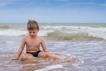 A little boy with a disgruntled, offended view sits in the water by the sea.