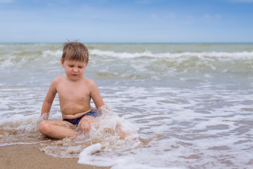 A little boy with a disgruntled, offended view sits in the water by the sea.