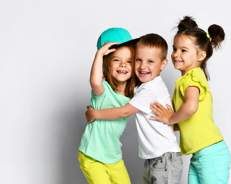 Studio portrait of children on a light background: full body shot of three children in bright clothes, two girls and one boy. Triplets, brother and sisters. hugging on camera. Family ties, friendship