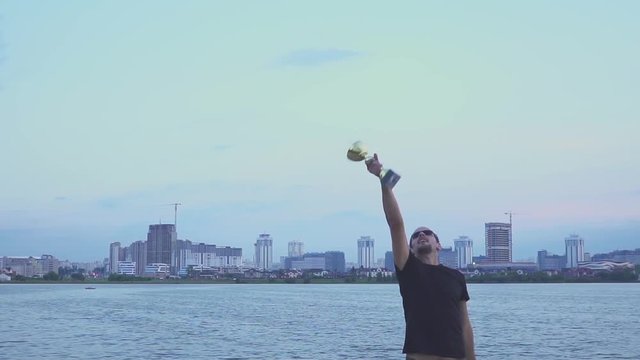 A young man with a cup in his hands rejoices in sports victory on background of the evening Minsk. Concept as victory, success and champion in sport. Slow motion.
