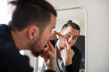 A young man indoors looking in the mirror, squeezing a pimple.