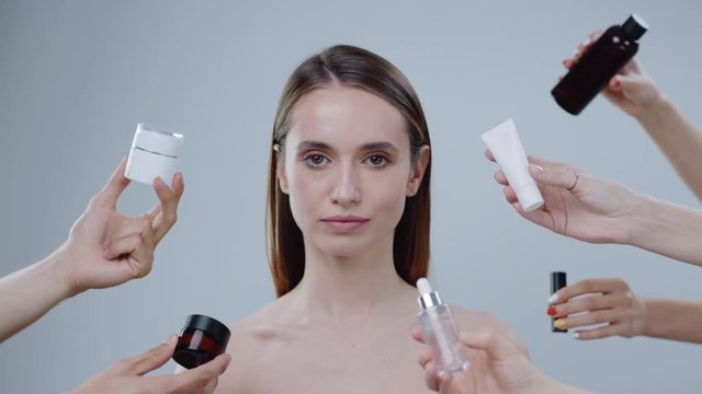 Close up of beautiful young woman look at camera on a grey background around a lot hands hold cosmetics appear and disappear attractive care clean complexion eyes face girl health makeup slow motion