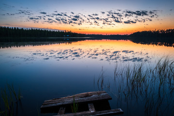 Colorful Sunset and loneliness at a remote finnish lake with beautiful reflections of clouds and dark boreal forest and old pier in foreground, Finland