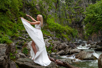 A young blonde girl in an elegant pose pulls up a boudoir dress in the mountains against a waterfall and stones raising her hands up like a ballerina on a warm summer day in Altai with green trees