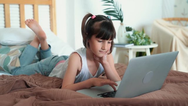 Little girl lying on bed, uses laptop, presses buttons on keyboard, slow motion