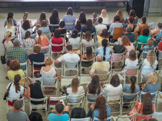 Many people sit indoors on light folding chairs. Rear view and top. The concept of the event - a seminar, lecture, training, training, watching a film, a play.