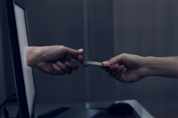 A hand that sends a credit card to a hand that stands out from a computer monitor in a dark tone.
